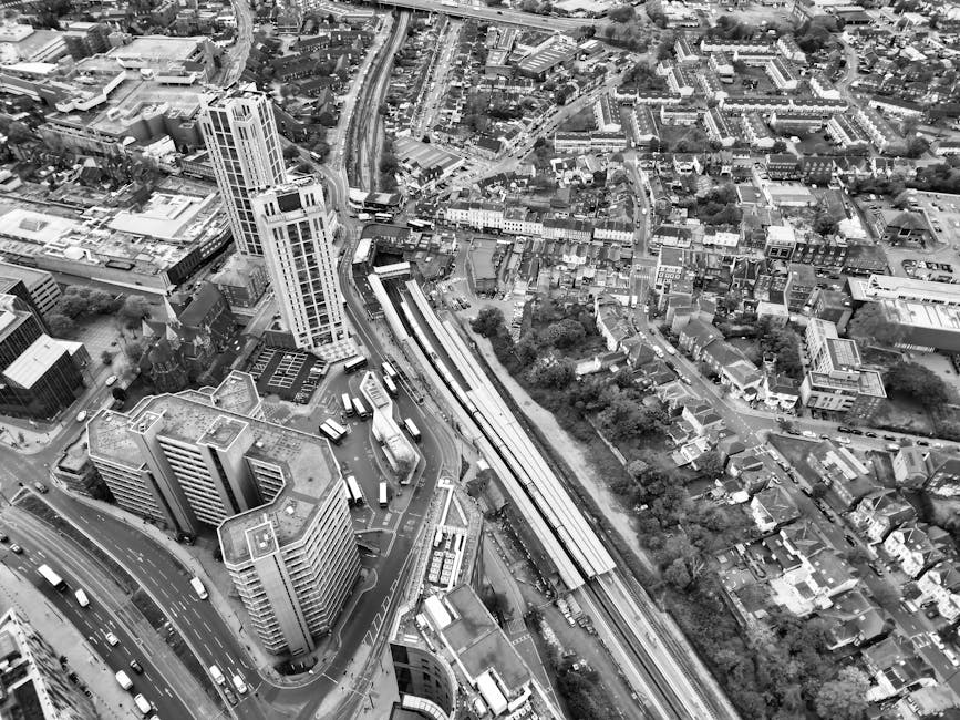 A black-and-white aerial photograph of an urban area showing a mix of high-rise office buildings and smaller residential houses. In the foreground, several multi-storey office blocks with flat roofs are visible, their facades featuring large windows. To the left, a curved road with multiple lanes is busy with vehicles, including buses and cars. In the middle of the image, a long, elevated railway line runs parallel to a canal or green corridor lined with trees, which separates the high-rise area from the smaller houses and residential streets in the background. The residential area consists of tightly packed houses with pitched roofs, narrow streets, and small gardens, extending to the upper right of the image. The overall scene depicts a densely built cityscape with prominent commercial buildings, transportation infrastructure, and surrounding residential neighborhoods, illustrating the scale and complexity of urban planning and waste collection requirements, as managed by waste clearance services like Waste Clearance Acton.