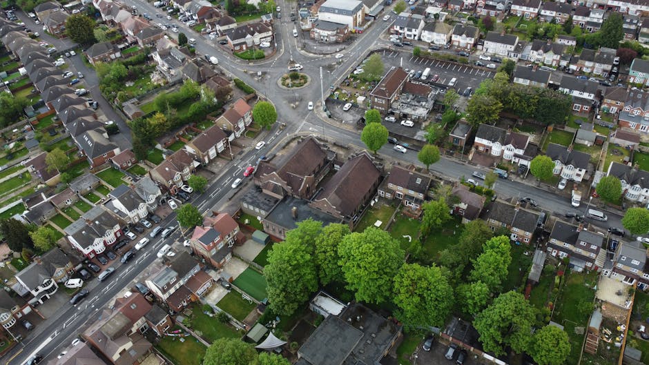 An aerial view of a residential neighbourhood showing a mix of detached, semi-detached, and terraced houses with predominantly brick exteriors and tiled roofs. The streets are lined with parked cars and small trees, with some green lawns and gardens visible in front and backyard spaces. A central roundabout with a small landscaped island features shrubs and grass, surrounded by a network of roads connecting the area. Larger buildings, possibly commercial or community facilities, are situated near the roundabout, with dark roofing and brick walls. Dense clusters of trees provide greenery throughout the area, including a few larger, leafy trees in backyards and along streets. The overall scene displays a typical suburban layout with a balance of residential housing, vegetation, and road infrastructure, suitable for interpreting the context of local rubbish removal or independent waste clearance services on-site.