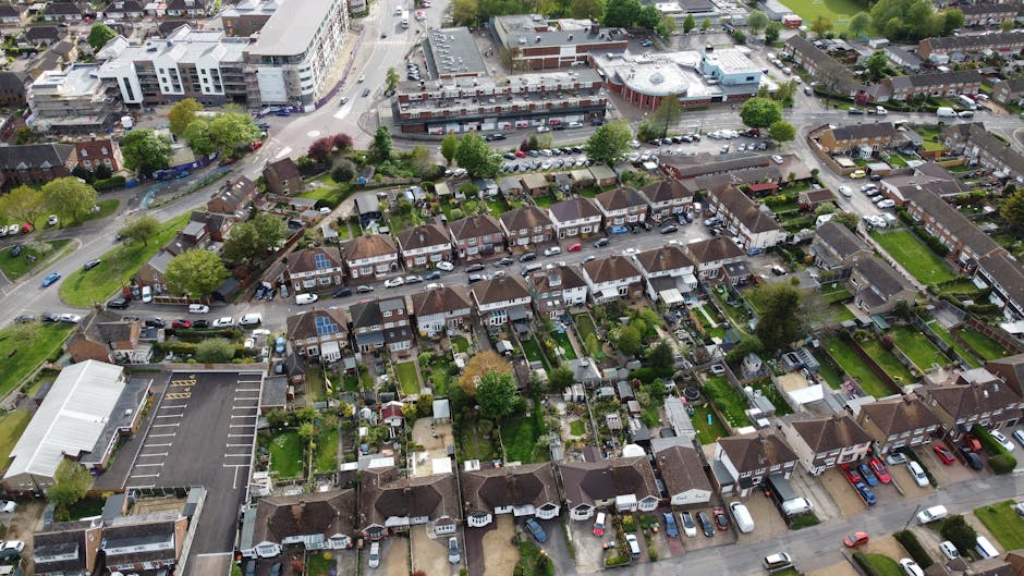 An aerial view of a residential neighbourhood showing a mix of detached, semi-detached, and terraced houses with predominantly brick exteriors and tiled roofs. The streets are lined with parked cars and small trees, with some green lawns and gardens visible in front and backyard spaces. A central roundabout with a small landscaped island features shrubs and grass, surrounded by a network of roads connecting the area. Larger buildings, possibly commercial or community facilities, are situated near the roundabout, with dark roofing and brick walls. Dense clusters of trees provide greenery throughout the area, including a few larger, leafy trees in backyards and along streets. The overall scene displays a typical suburban layout with a balance of residential housing, vegetation, and road infrastructure, suitable for interpreting the context of local rubbish removal or independent waste clearance services on-site.