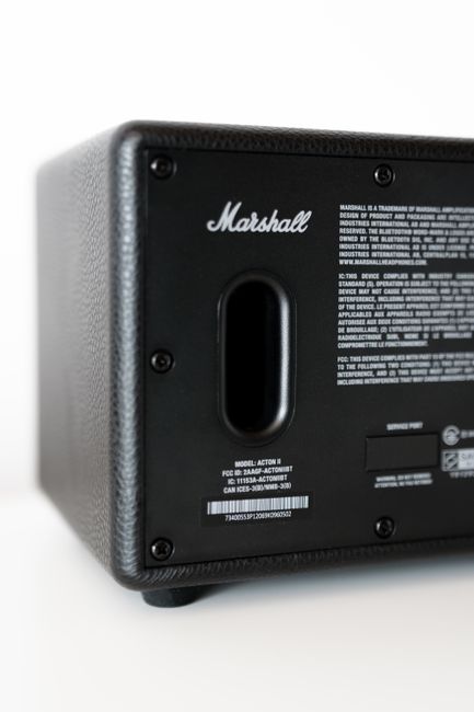 Close-up image of the underside of a black Marshall speaker with a textured finish, showing a central oval port and several visible screws securing the casing. The background is plain white, highlighting the speaker's rectangular shape and corner details. This photograph emphasizes the material and construction of the speaker, which is typical for audio equipment often managed or cleared in waste removal services. Though primarily focused on the device, the image relates to bulk or electronic waste collection, as offered by waste clearance services like Waste Clearance Acton, in contexts involving proper disposal of electronic and audio equipment. The lighting is soft and even, clearly revealing the textured surface and finish of the speaker, suggesting a professional and neutral aesthetic in line with accurate waste management depiction. Overall, the image effectively supports the context of handling electronic waste as part of a comprehensive rubbish removal process.