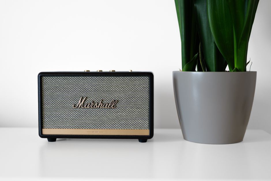 A compact Marshall portable speaker with a black casing, beige woven grille, and metallic logo positioned on the front, situated on a white surface against a plain white background. To the right of the speaker is a modern, rounded grey pot containing a green plant with broad, glossy leaves. The speaker's textured grille and sleek finish contrast with the smooth, matte surface of the plant pot, creating a simple yet functional scene. The setting appears indoors, with natural or neutral lighting illuminating the objects clearly. This image reflects a clean, organized environment where electronic gadgets and decorative indoor plants coexist, resonating with the concept of maintaining tidy and clutter-free spaces, which can relate subtly to the services offered by waste clearance and rubbish removal companies like Waste Clearance Acton, especially when cleaning or clearing out personal or domestic spaces. The overall composition emphasizes minimalism and practical objects that represent a typical complementary household environment, relevant in contexts of private waste handling or on-site clearance activities. The detailed description captures all visible elements clearly and neutrally, suitable for accessibility and SEO purposes.